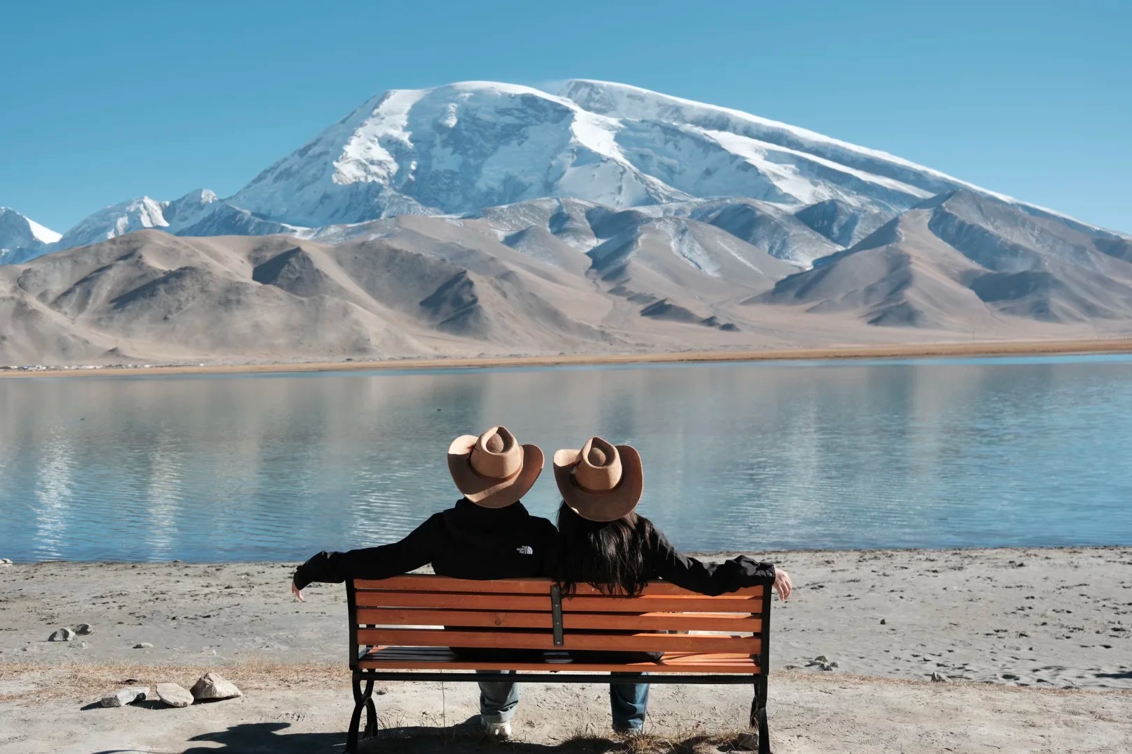 Cặp đôi tạo dáng trước mặt hồ, phía sau là núi tuyết và trời xanh.