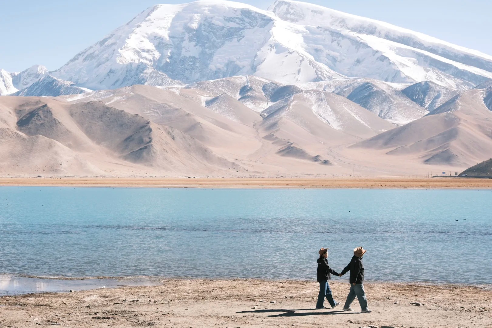 Cặp đôi tạo dáng trước mặt hồ, phía sau là núi tuyết và trời xanh.